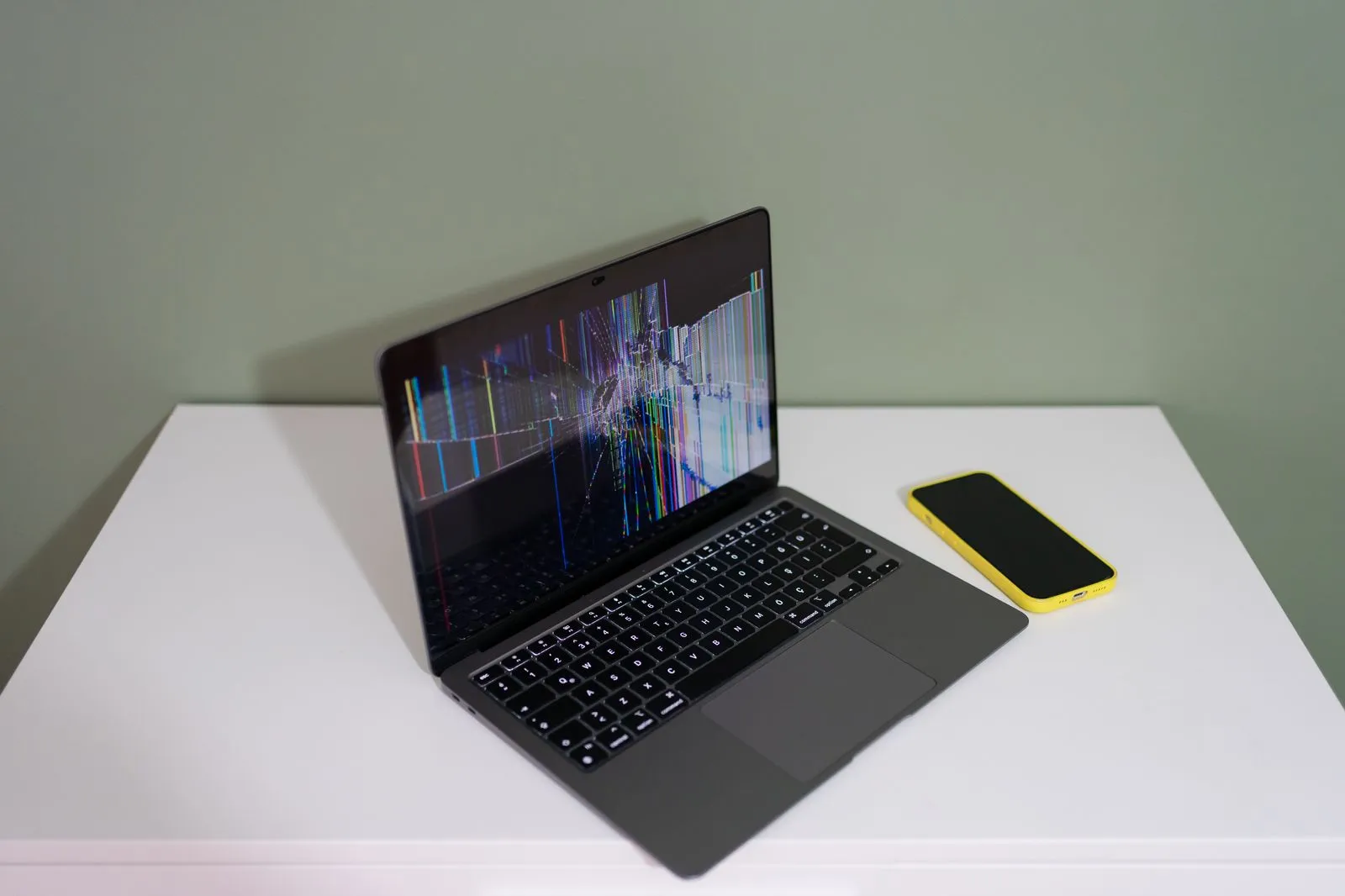 A damaged laptop screen with colorful lines and a yellow smartphone on a white desk.